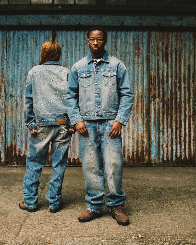 Two models wearing light-wash blue denim jackets and jeans stand in front of a weathered blue corrugated metal wall. One model faces forward wearing a denim jacket and jeans, while the other stands behind facing away.